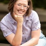 Picture of person in purple shirt sitting with head in hand looking ahead and smiling.