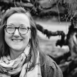 Gray scale photograph has Chessie with a smile, scarf, glasses, with the backdrop of a Japanese Maple tree covered in moss on the branches