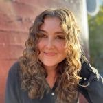 Curly-haired white woman with a soft smile, wearing a black shirt in front of a red brick wall.